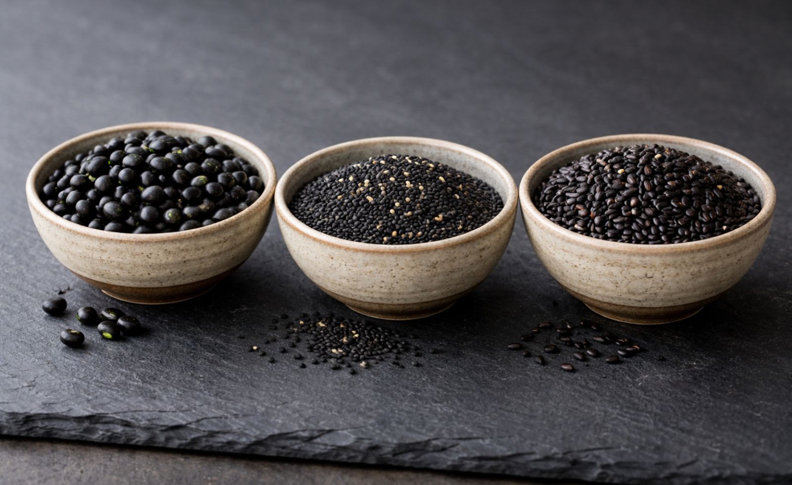 Trois petits bols en céramique écrus disposés côte à côte contenant respectivement des haricots noirs, du sésame noir et du riz noir